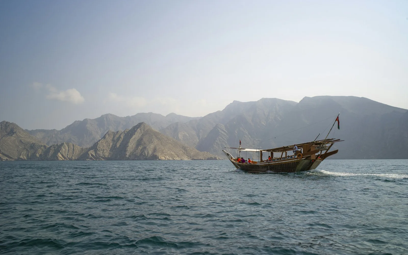 A traditional wooden dhow on turquoise water below steep limestone fjord cliffs in Musandam